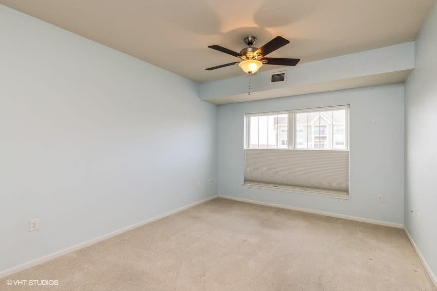 An empty room with light blue walls, featuring a ceiling fan and a window with blinds.