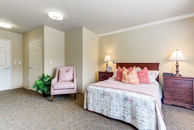 A cozy bedroom with a neatly made bed, a pink chair, and a bedside lamp.