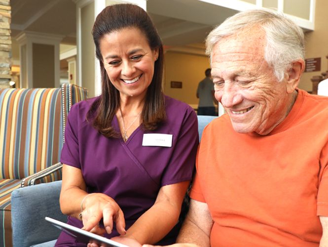 A caregiver assisting a senior man with a tablet, both are smiling happily.