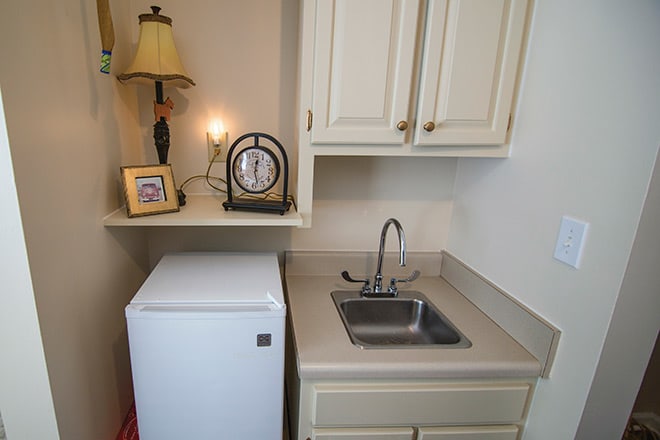 A small kitchen area with a sink, mini fridge, and overhead cabinets.
