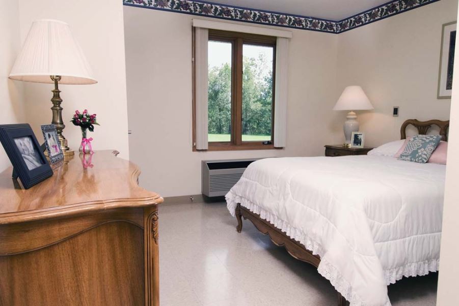 A cozy bedroom with a white bedspread, wooden furniture, and natural light from the window.