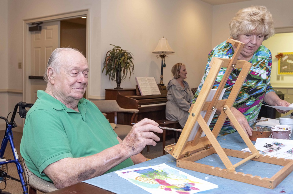 A group of seniors engaging in a painting activity indoors, with one man working on an artwork using an easel.