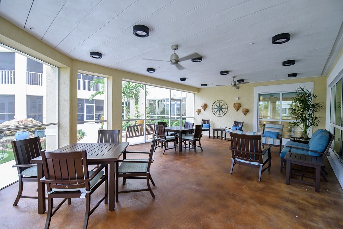 A cozy enclosed patio with multiple tables and chairs arranged for seating, surrounded by large windows and featuring ceiling fans overhead.