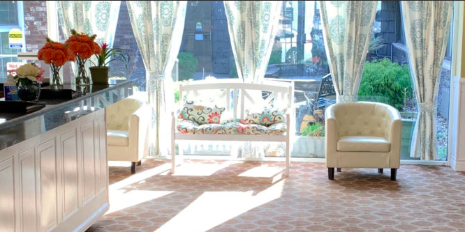 A sunlit room with two chairs, a decorative bench, and large windows with sheer curtains.
