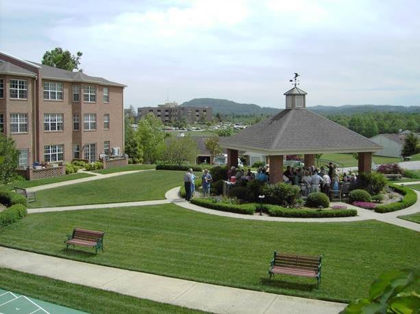 A scenic view of a well-maintained lawn with a gazebo and benches, surrounded by buildings and a group of people gathered nearby.