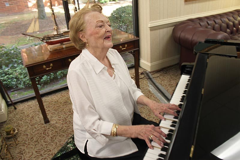 An elderly woman playing the piano indoors, wearing a white shirt and bracelets.
