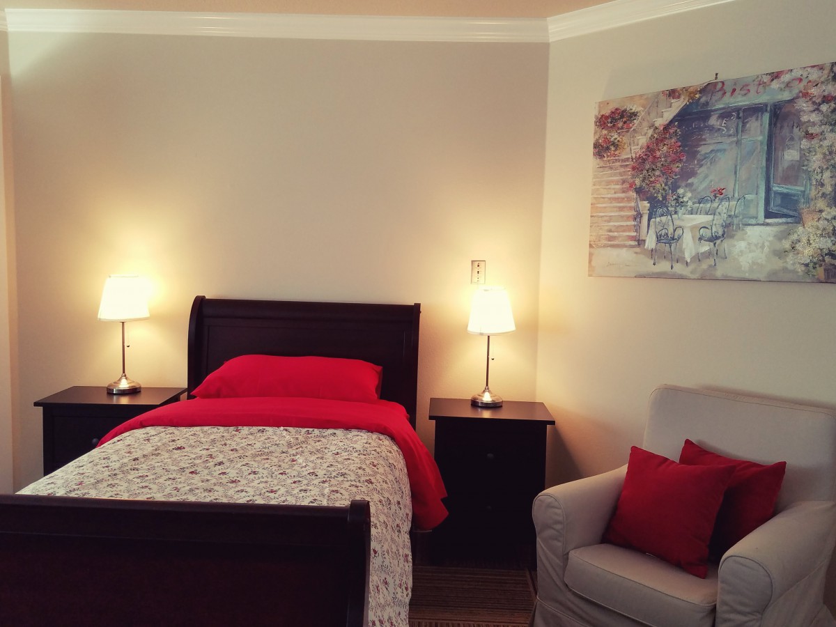 A cozy bedroom featuring a single bed with red bedding, flanked by nightstands and lamps, alongside an armchair with matching red cushions.