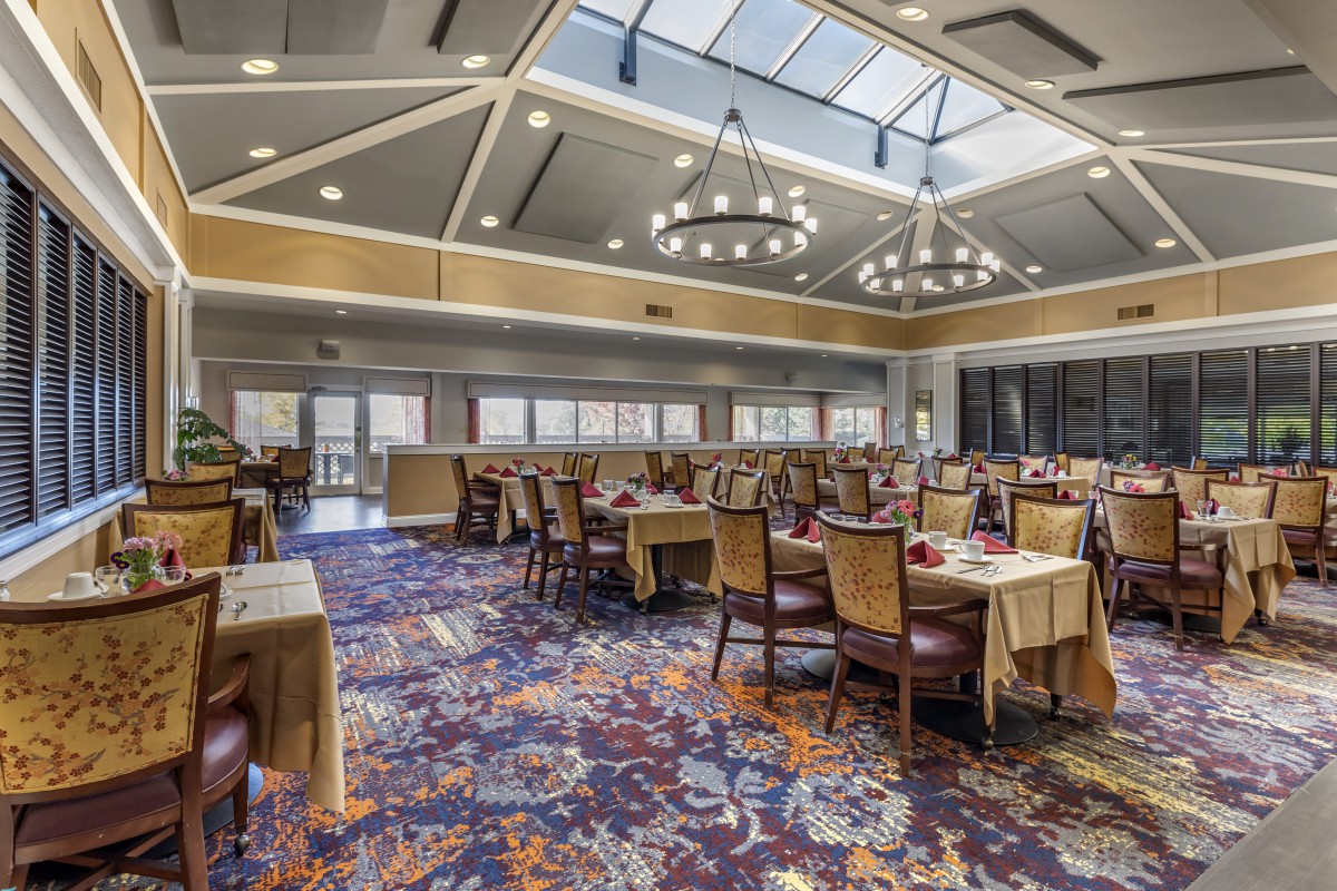 A spacious dining room with elegant chandeliers and a skylight, featuring multiple tables and chairs arranged on a colorful carpet.