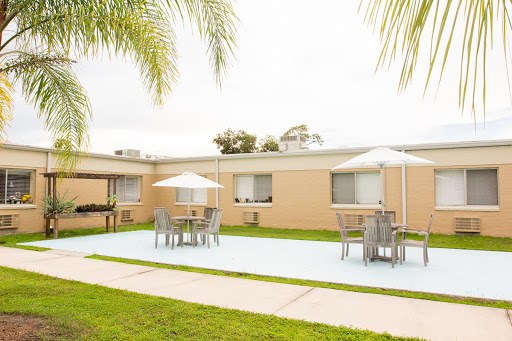 A serene outdoor courtyard featuring tables and chairs under umbrellas, surrounded by a well-maintained lawn and palm trees.
