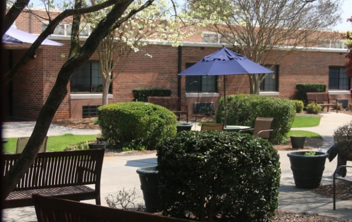 A peaceful courtyard with benches and a blue umbrella, surrounded by manicured bushes and trees.