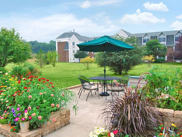A serene garden patio with a table and chairs, shaded by an umbrella, surrounded by lush greenery and colorful flowers.