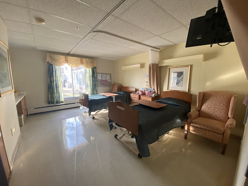 A well-lit hospital room featuring two patient beds, chairs, and a window with a view.