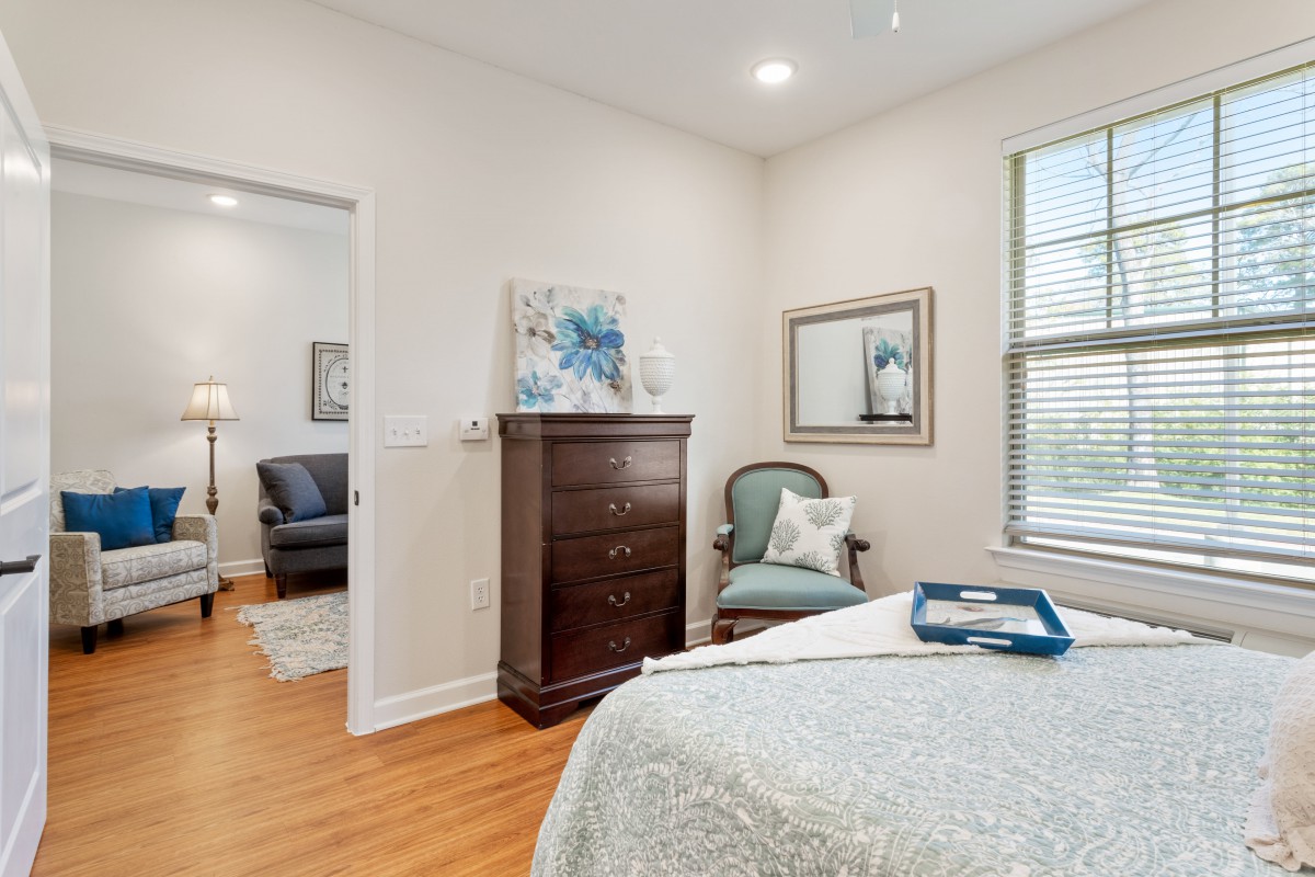 A cozy bedroom featuring a wooden dresser, an upholstered chair, and a neatly made bed with soft lighting.
