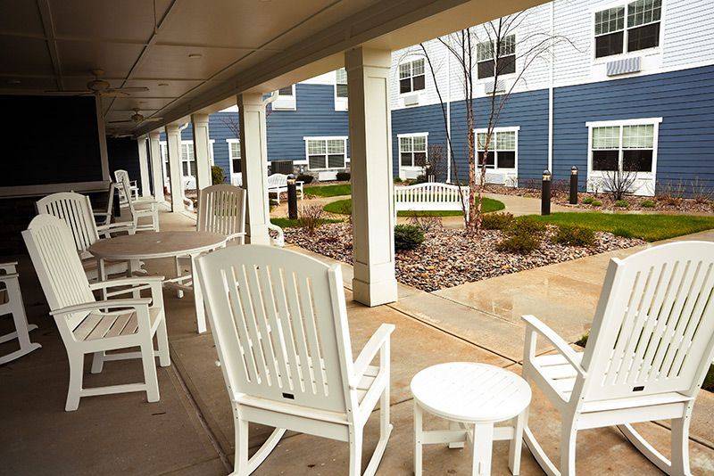 A covered patio area with white rocking chairs and small tables overlooking a landscaped courtyard in front of a building.