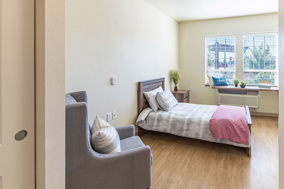 A minimalist bedroom with a neatly made bed, an armchair, and natural lighting through a window.