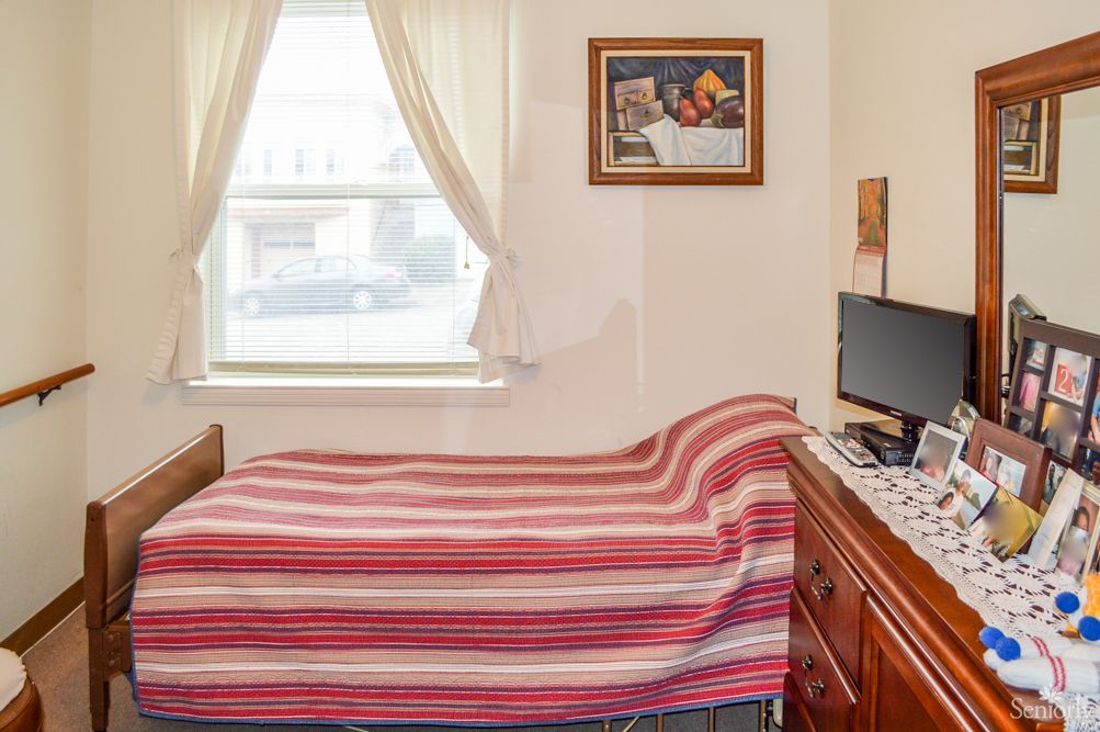 A cozy bedroom with a neatly made bed, a computer on the desk, and light filtering through curtained windows.