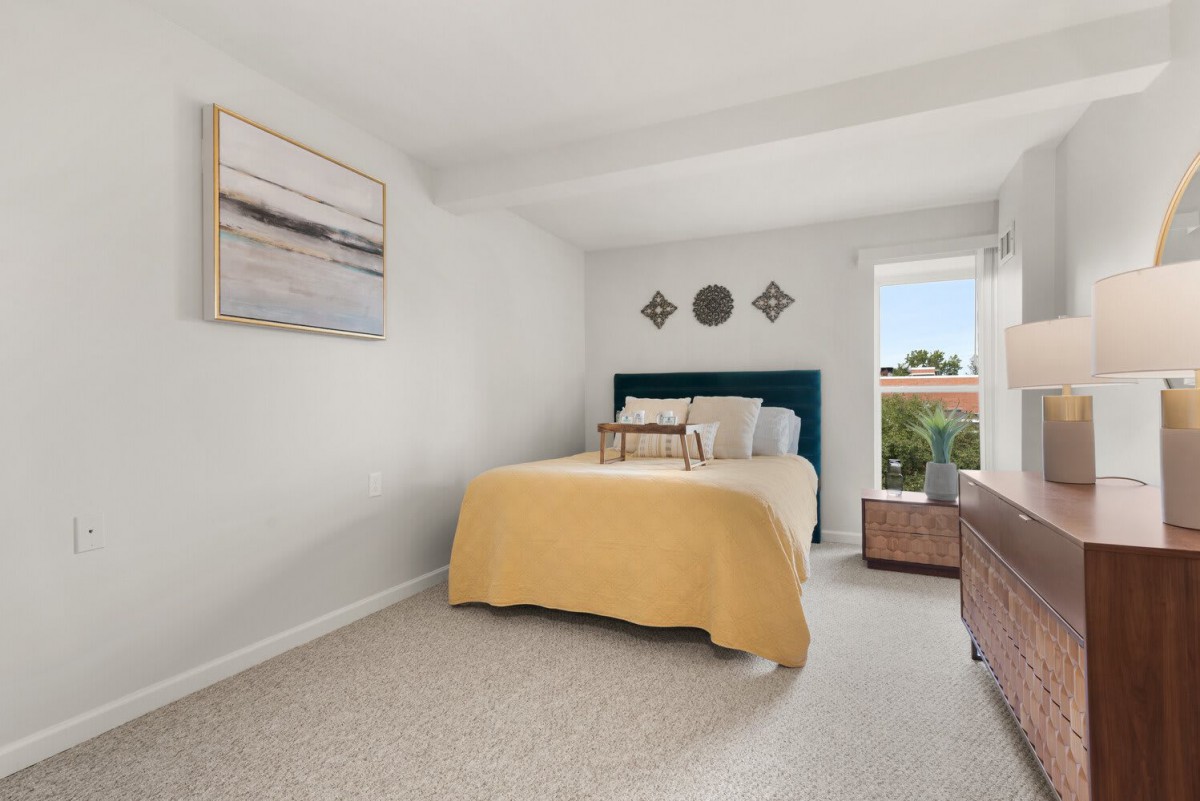 A cozy bedroom with a neatly made bed featuring a yellow blanket, wall decorations, and a window providing natural light.