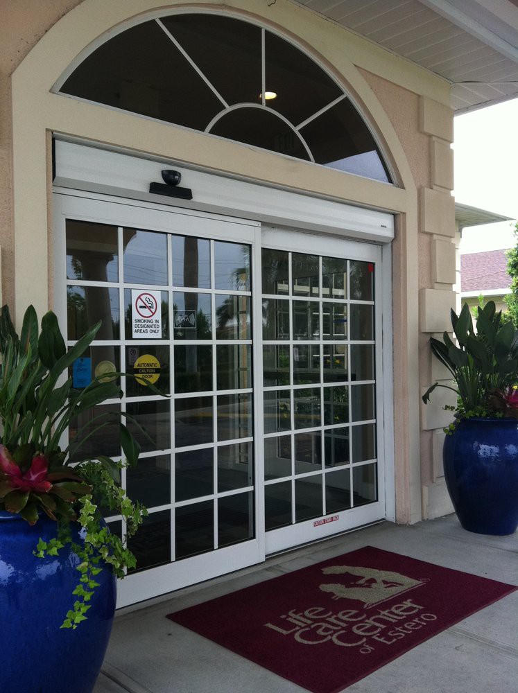 A building entrance featuring a sliding glass door flanked by large decorative blue pots with vibrant plants.