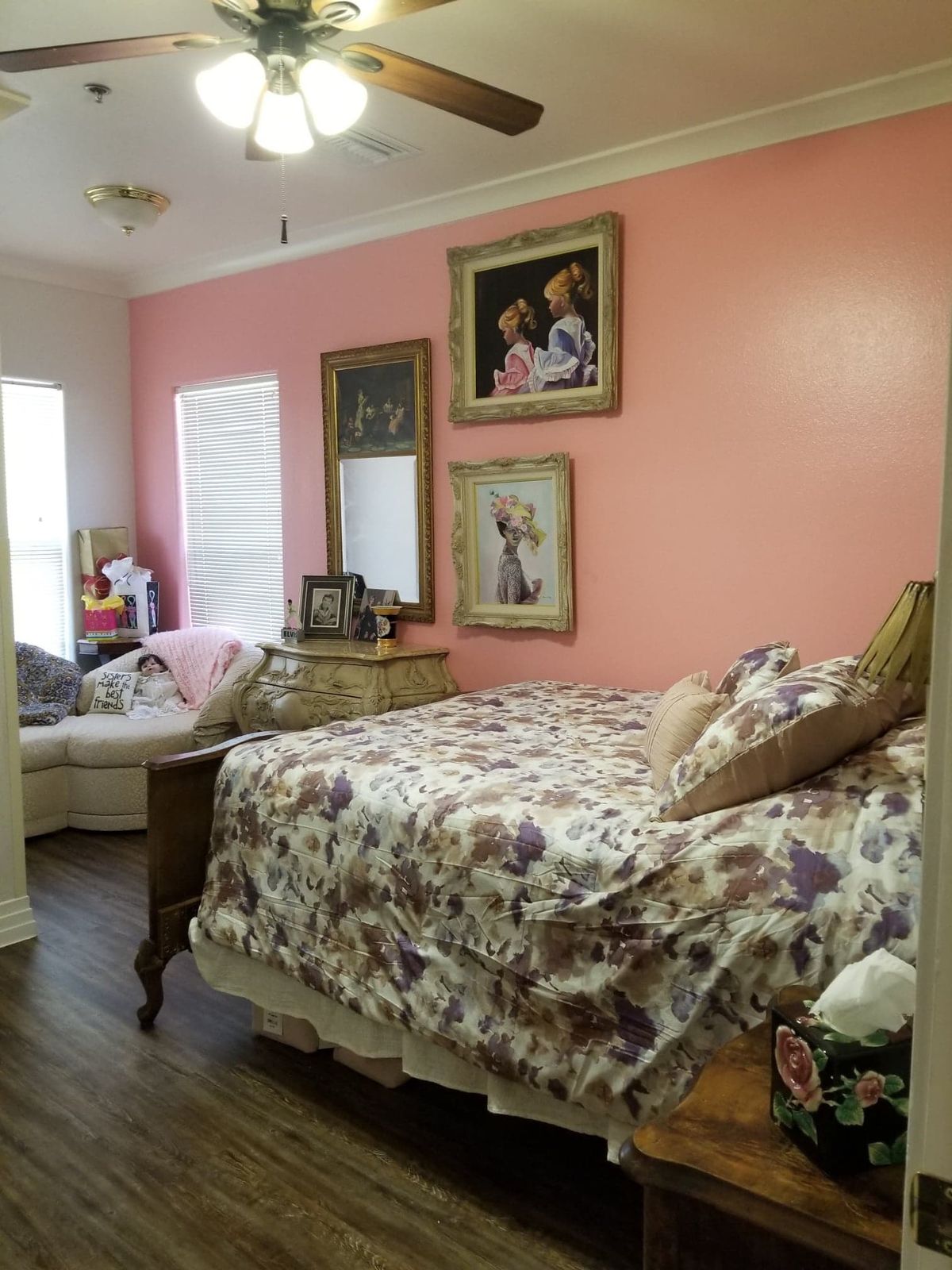 A cozy bedroom with a pink accent wall and floral bedding, featuring decorative paintings and wood flooring.