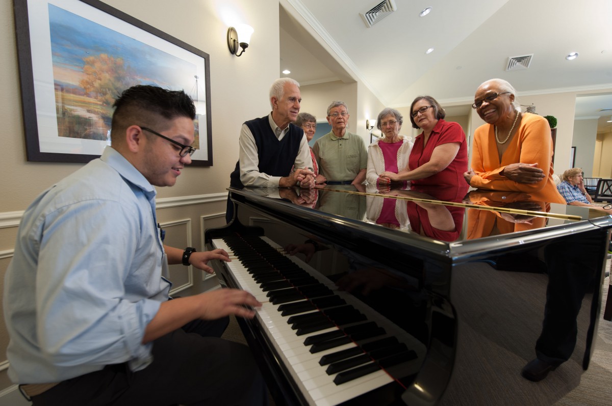 A pianist plays for an audience gathered around a piano indoors.