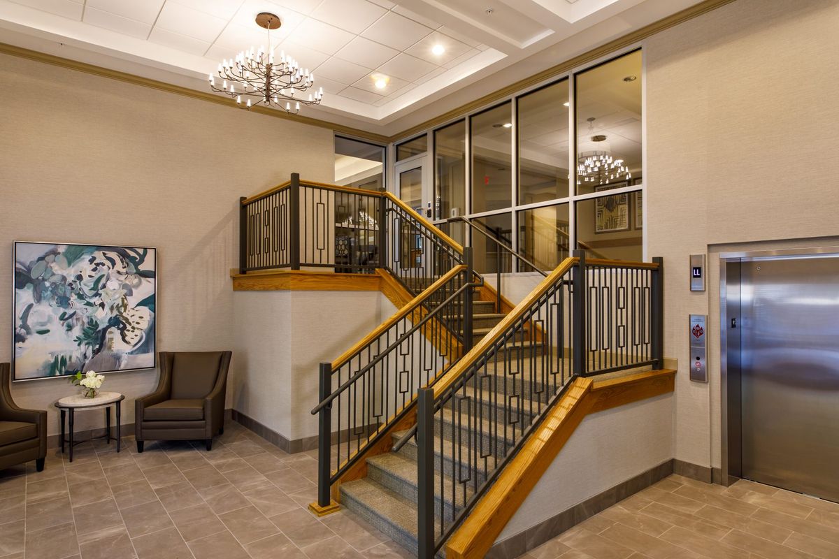 A modern lobby interior featuring a staircase with wooden railings, a seating area, and chandeliers.