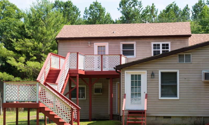 A two-story house with an exterior staircase leading to a balcony, surrounded by trees.