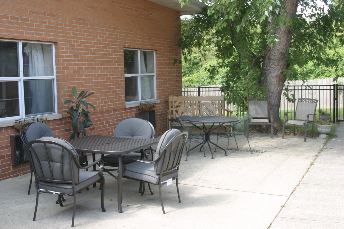 A tranquil outdoor patio area with tables and chairs beside a brick building, shaded by trees.