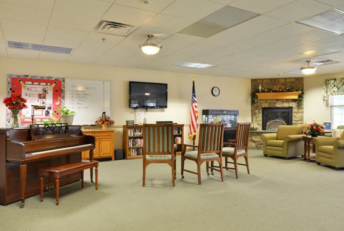 A community room with arranged seating, a piano, and an American flag, featuring a television and fireplace.