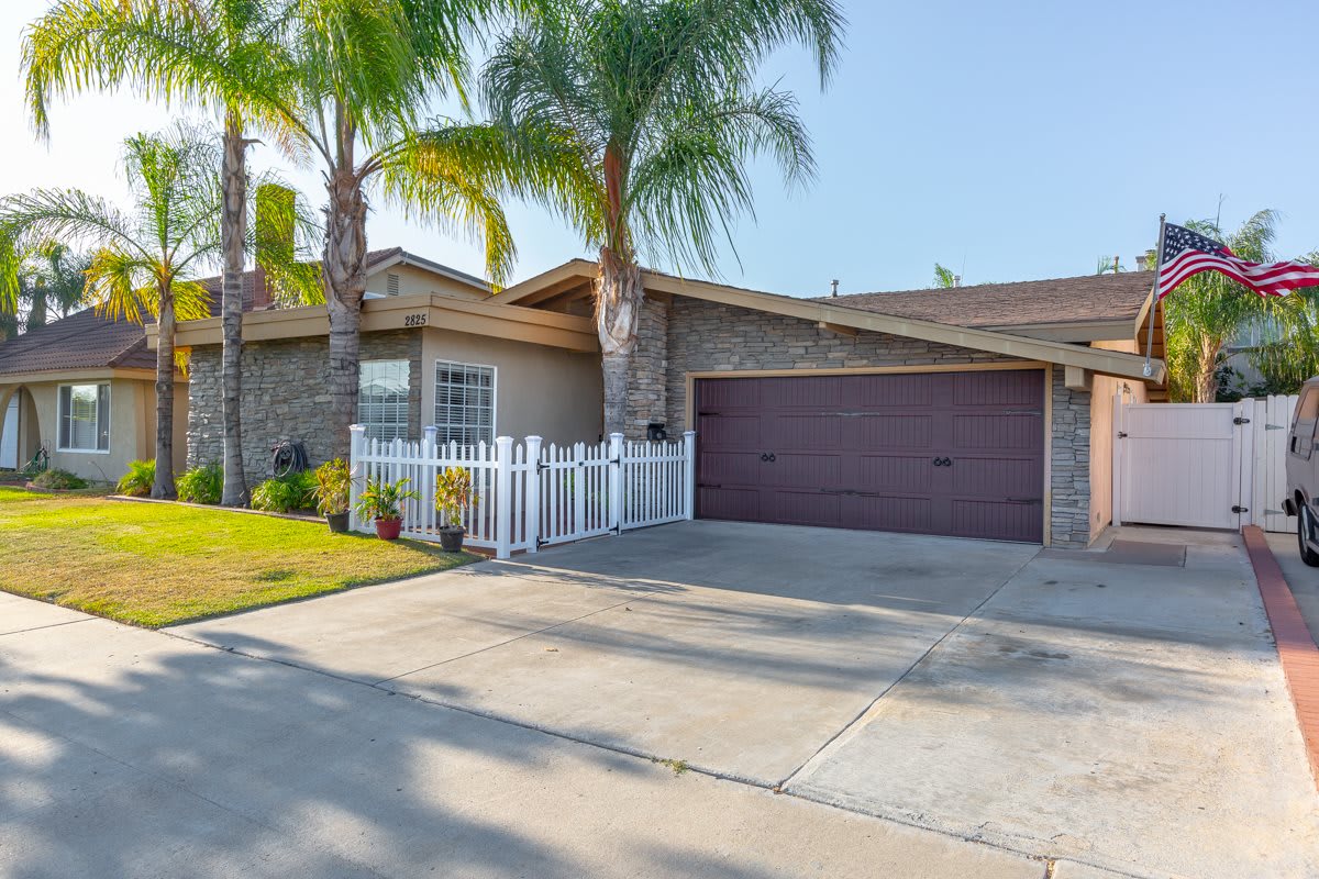 A suburban house with a driveway and garage, surrounded by palm trees and a white fence.