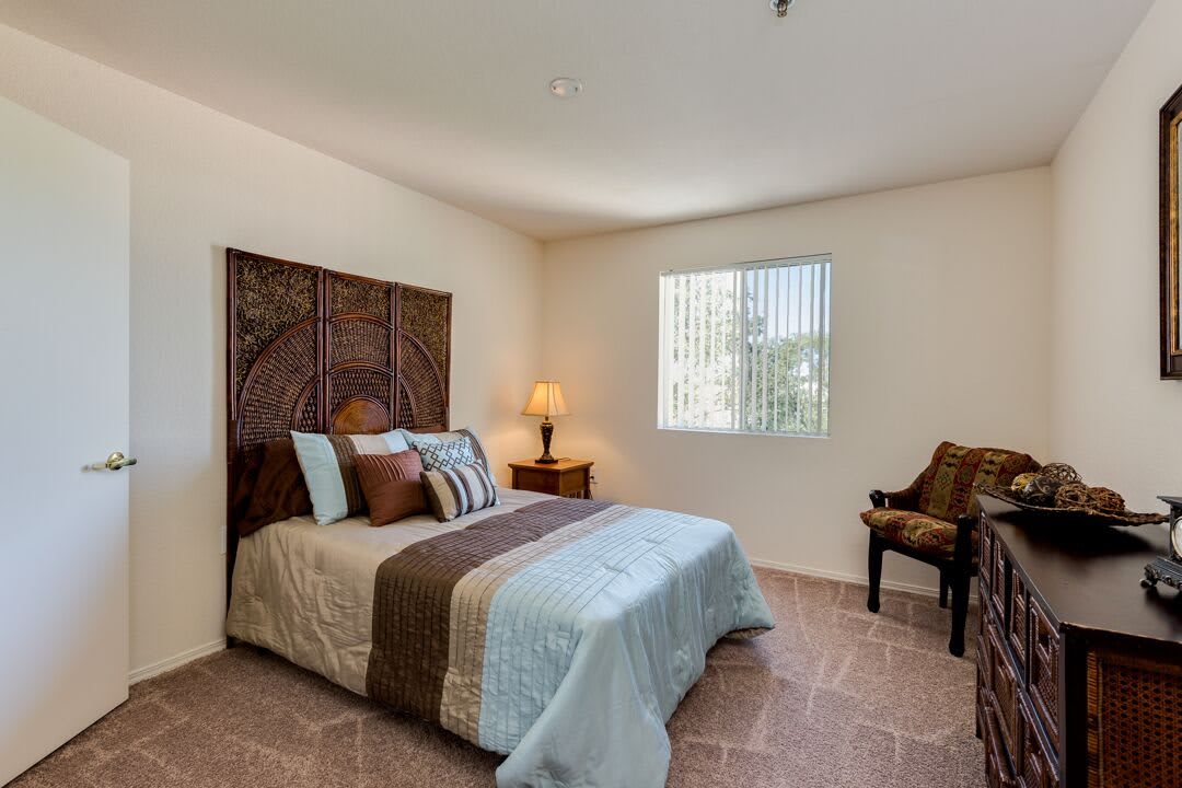 A cozy bedroom with a bed, side table, lamp, and chair next to a window.