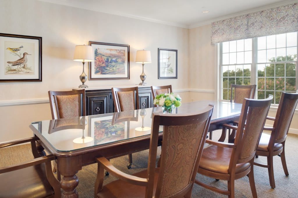 A well-lit dining room with a glass-topped table, wooden chairs, and decorative elements including artwork and a flower arrangement.