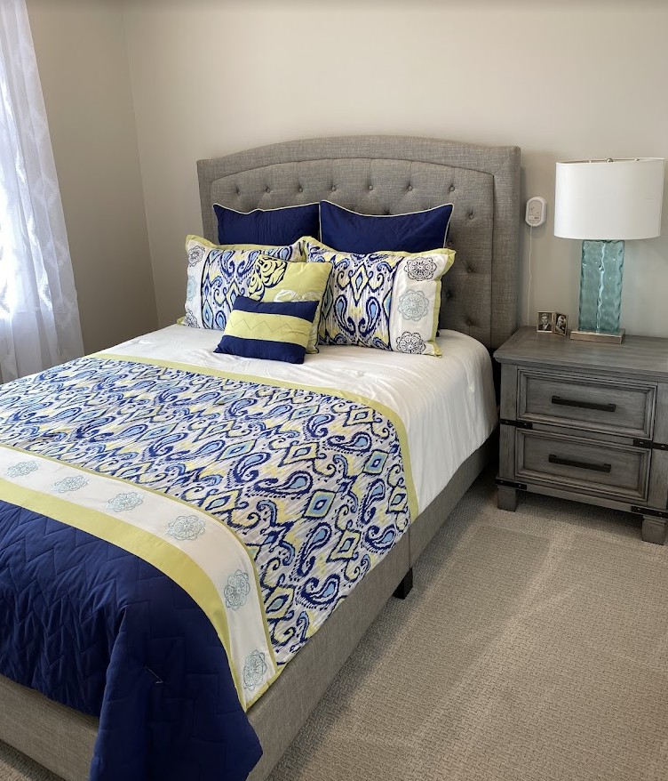 A well-decorated bedroom with a bed adorned with blue, yellow, and white patterned bedding and pillows, alongside a nightstand and lamp.