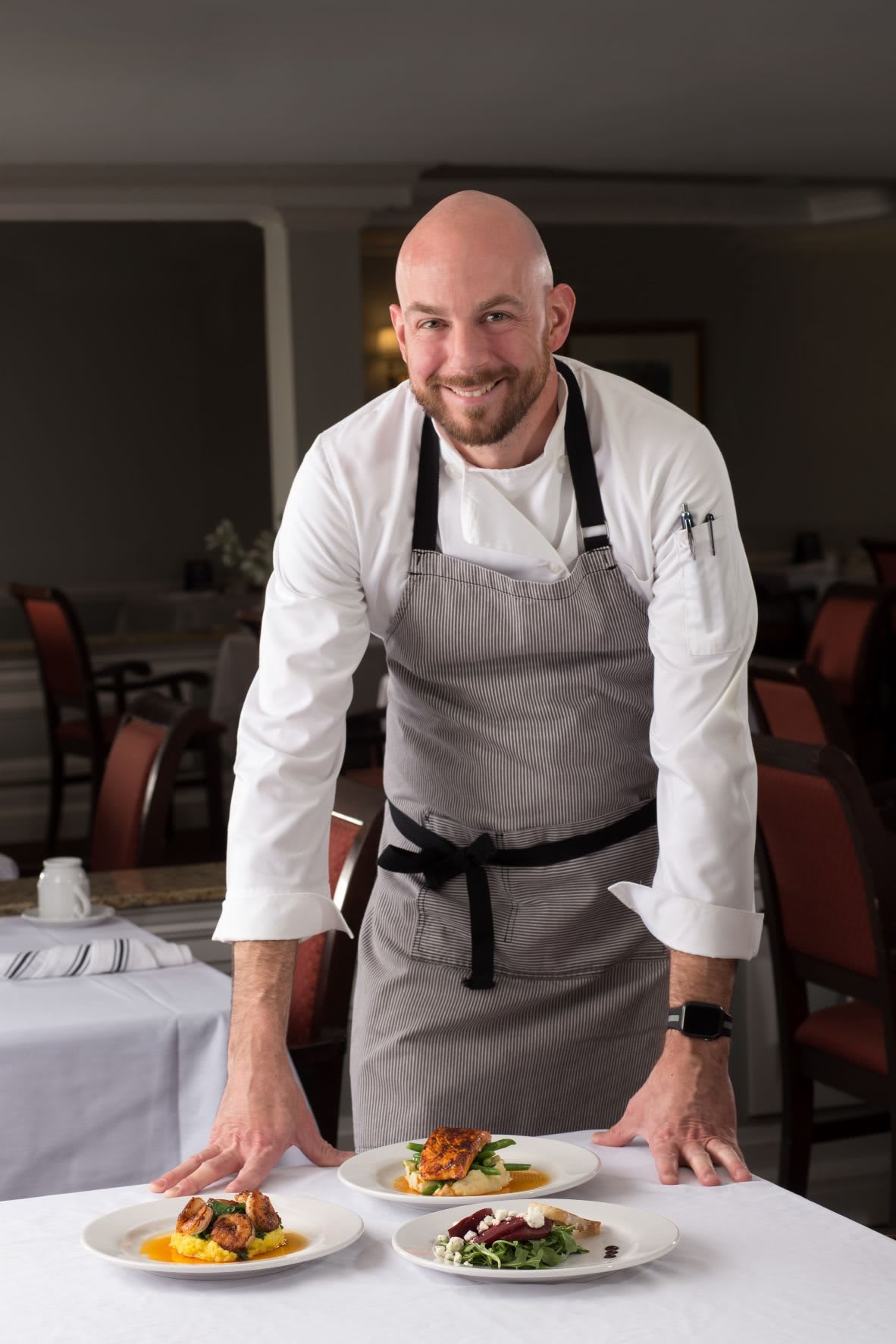A chef standing in a restaurant setting, wearing professional attire and an apron.