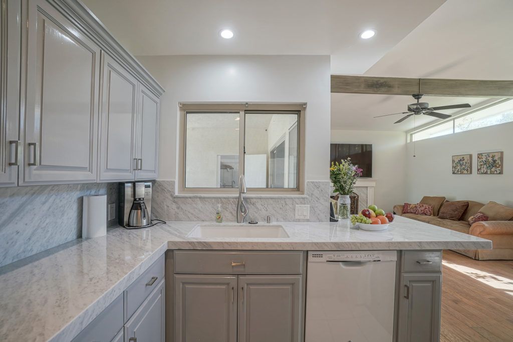 A modern kitchen interior with neutral tones, featuring sleek cabinets, a countertop with a fruit bowl, and a view into the adjacent living area.