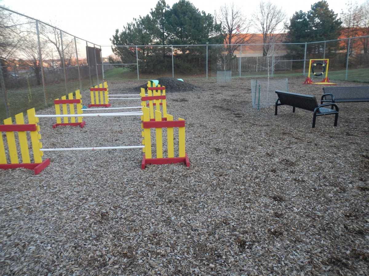 A fenced-off dog park featuring an agility course with hurdles and gravel ground.
