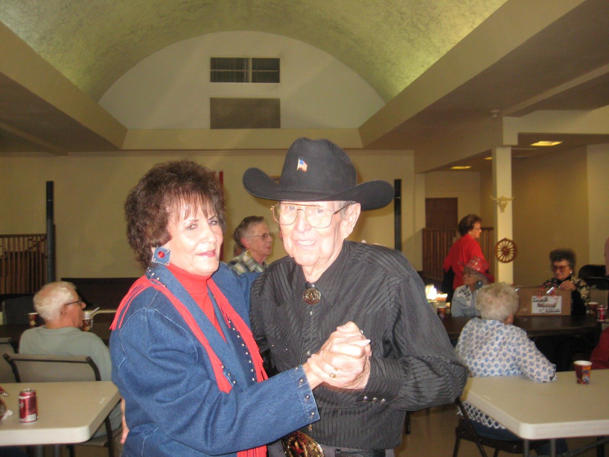 An elderly couple is dancing together, with the man wearing a cowboy hat, in an indoor setting.