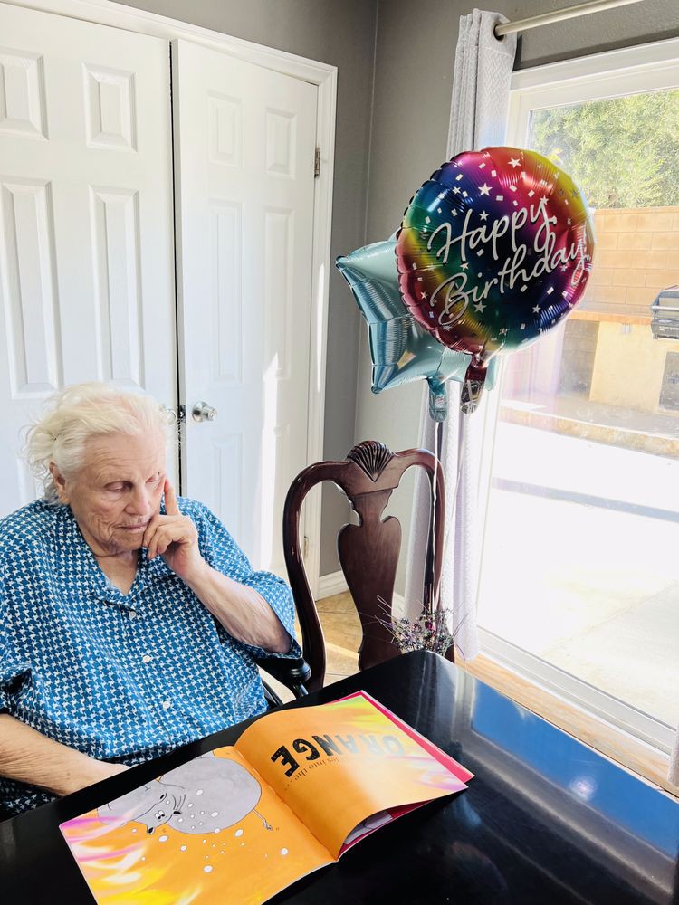 An elderly woman sitting at a table with birthday balloons and a card, celebrating her special day.