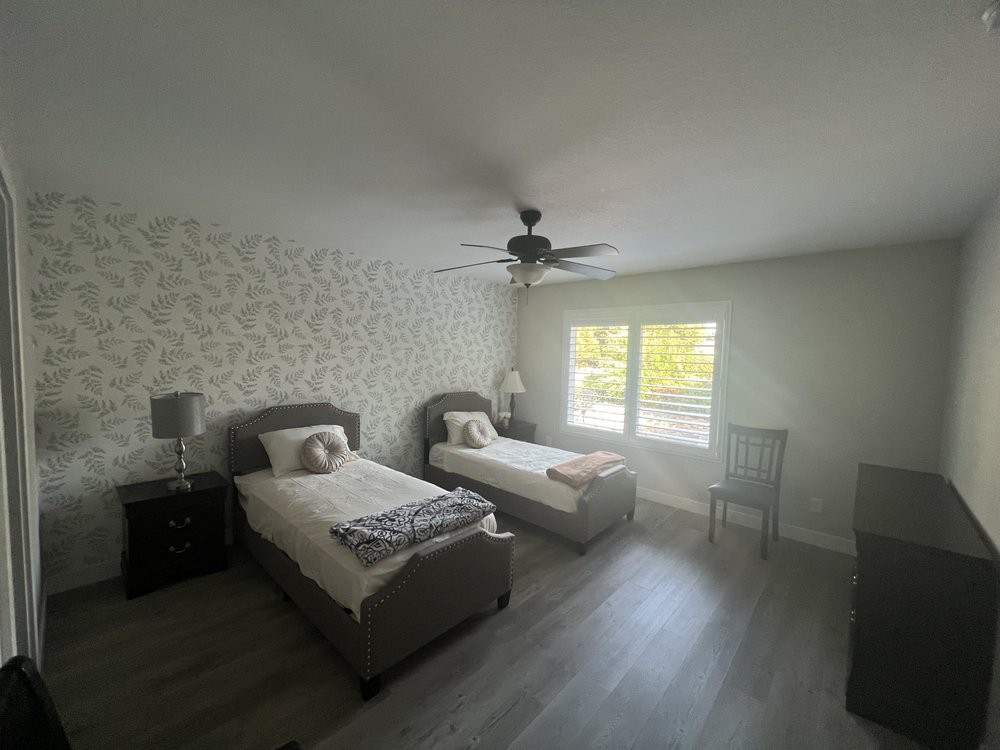 A bright bedroom featuring twin beds, a patterned wallpaper accent wall, nightstands with lamps, a window letting in natural light, and a ceiling fan.