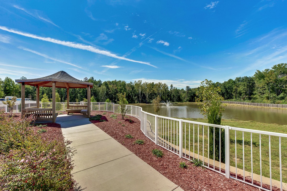 A scenic pathway leading to a gazebo by a pond, surrounded by lush trees under a clear blue sky.