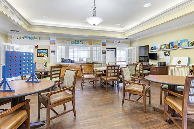 A spacious and well-lit community room with wooden floors, featuring several tables and chairs arranged for social gatherings.