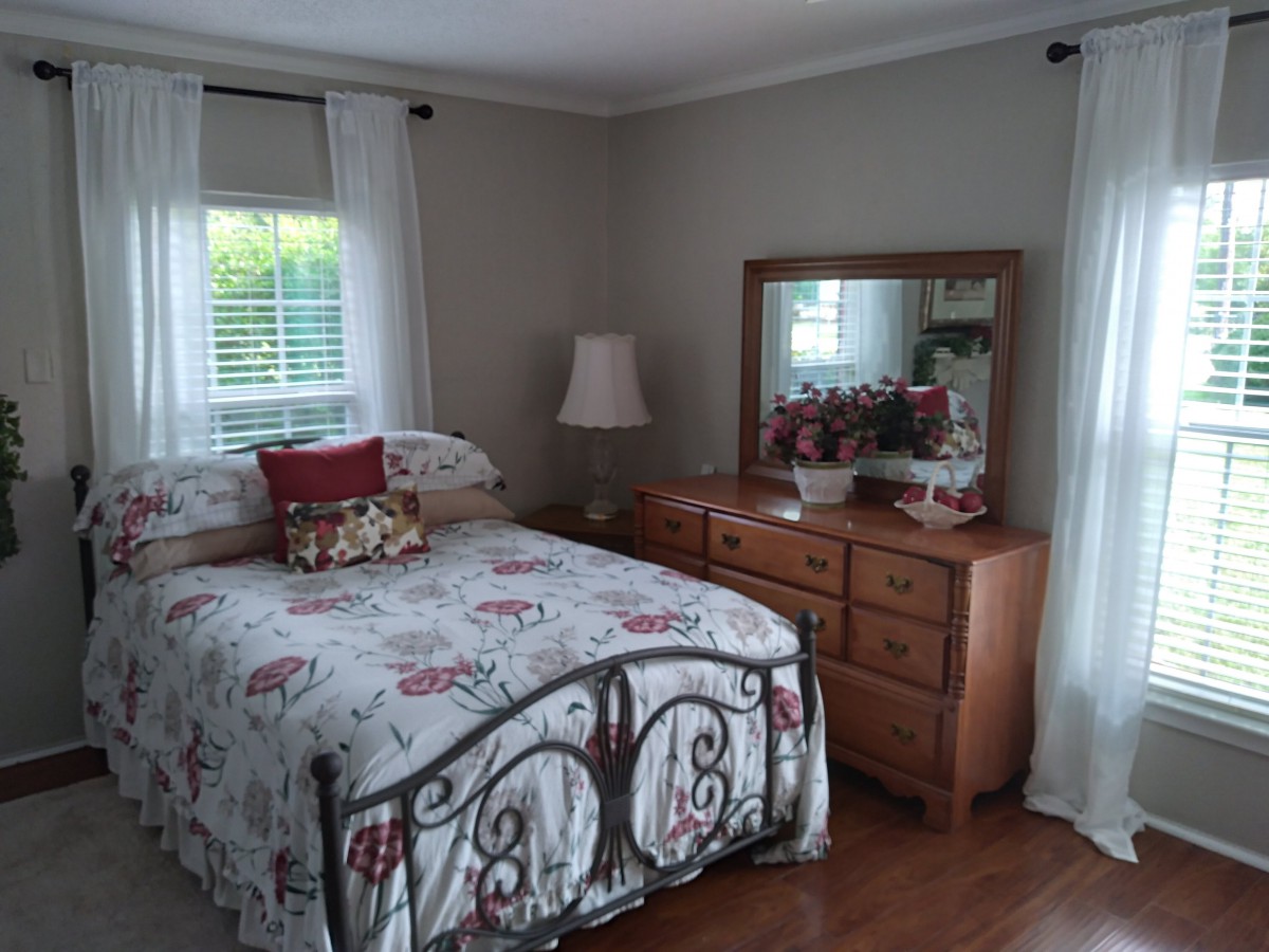 A cozy bedroom with a floral-patterned bed, a wooden dresser with a mirror, and soft natural light coming through the window.