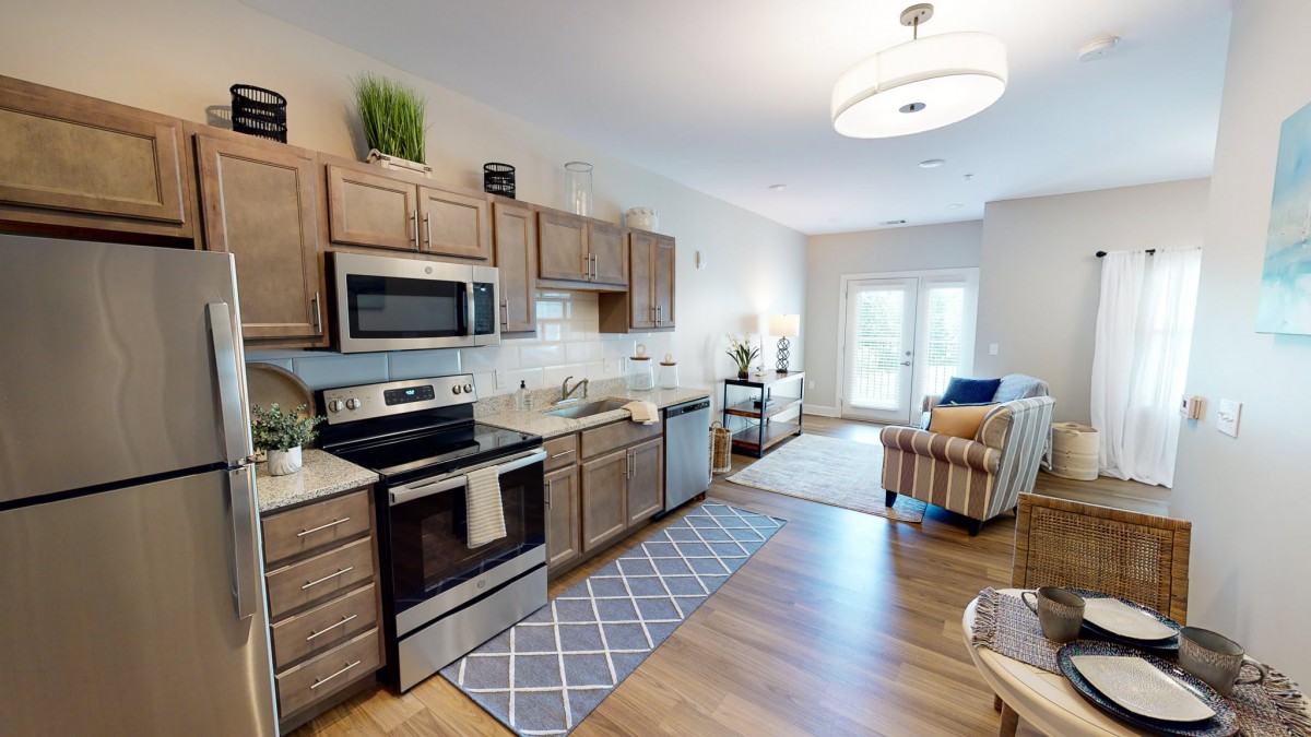 A modern open-concept kitchen with wooden cabinets and stainless steel appliances, leading into a cozy living area.