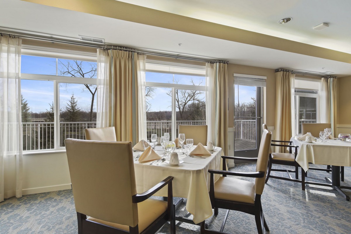 A well-lit dining room with elegantly set tables, overlooking a scenic view through large windows.