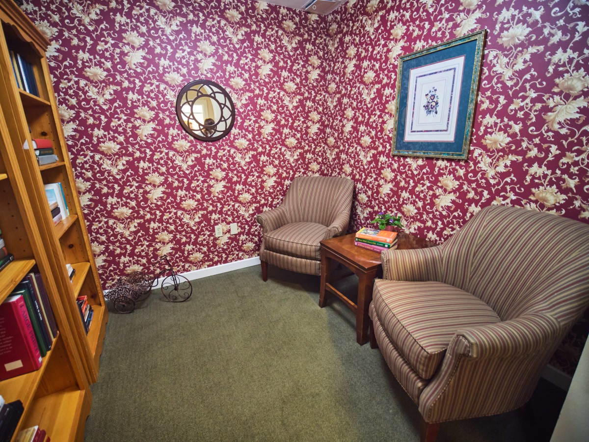 A cozy living room with vintage wallpaper, two armchairs, a small table, and a bookshelf.