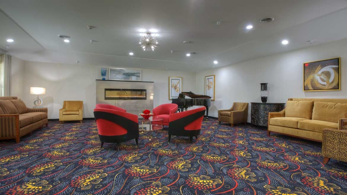 A modern lounge area featuring red chairs, a piano, and a colorful carpet.