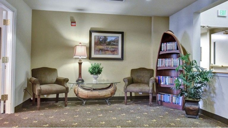 A cozy waiting area with two chairs, a glass top table, a lamp, and a bookshelf filled with books.