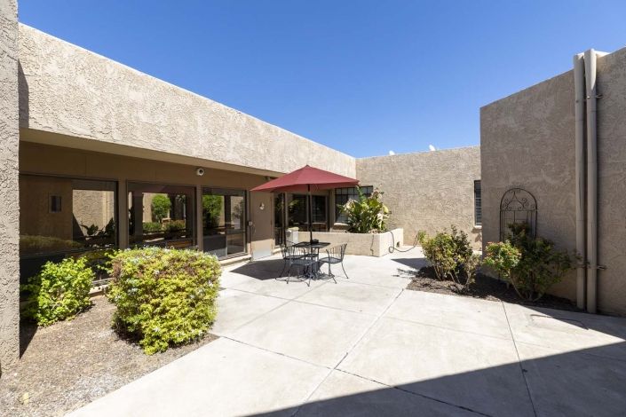 A sunny courtyard featuring a patio with a red umbrella, table, and chairs surrounded by desert plants.