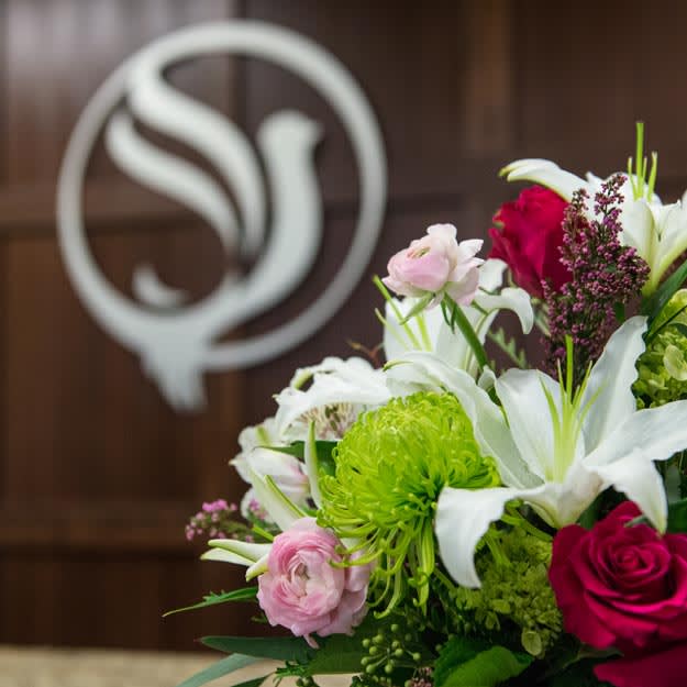 A vibrant bouquet of flowers including lilies and roses, set against a blurred logo background.