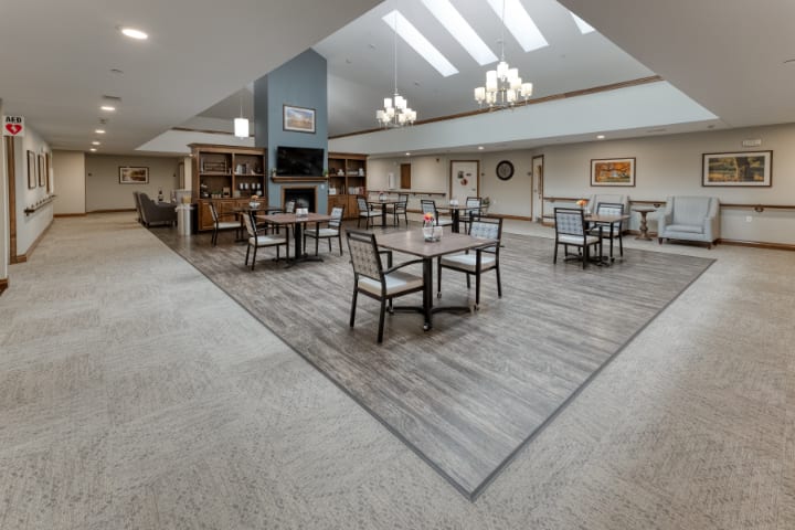 A spacious dining area with tables and chairs, featuring a central fireplace and overhead lighting under a vaulted ceiling.