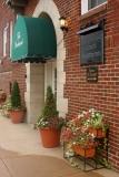 A building entrance with a green awning and potted plants, featuring a brick exterior.